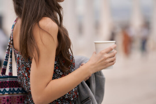 Unrecognizable Young woman holding a coffee cup on a busy street, focus on hand with blurred background.