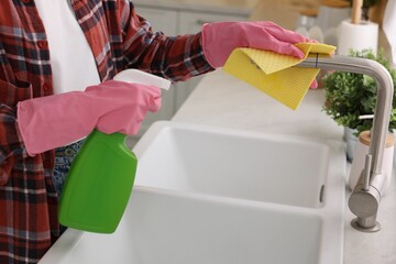Woman with spray bottle and microfiber cloth cleaning water tap in kitchen, closeup