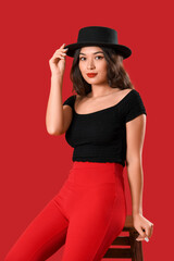 Beautiful young Asian woman in stylish outfit sitting on chair against red background