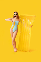 Surprised young woman in tank swimsuit and with inflatable mattress on yellow background