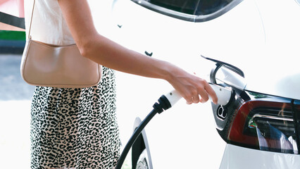 Young woman travel with EV electric car charging in green sustainable city outdoor garden in...