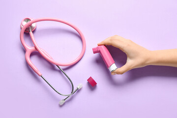 Child's hand with asthma inhaler and stethoscope on lilac background