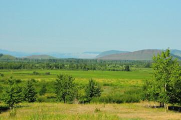 View of the clearing at the edge of the forest on a sunny day and a range of hills with fog on the tops.