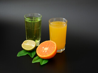 Two tall glasses with different citrus juices on a black background, next to the halves of ripe orange and lime with leaves.