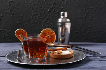 Metal plate with glasses of tasty Old Fashioned Cocktail, dried orange slices and bowl of sugar on wooden table near black wall