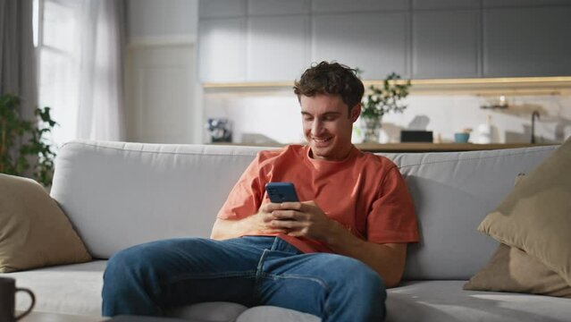 Cheerful man chatting smartphone cozy sofa. Happy smiling guy creating message