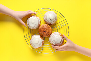 Female hands with cooling rack and cinnamon buns on yellow background