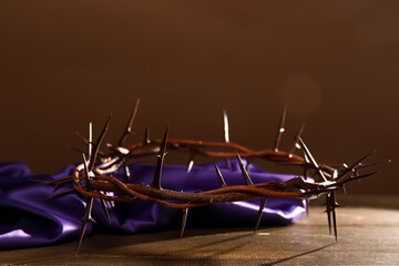 Crown of thorns with purple fabric on wooden table against brown background