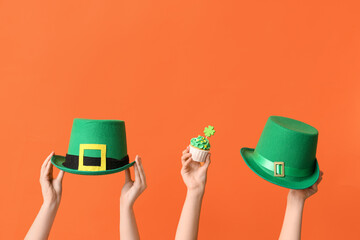 Female hands with tasty cupcake and leprechaun hats for St. Patrick's Day on orange background