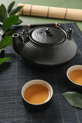 Bamboo mat with cups of tea and teapot on green background