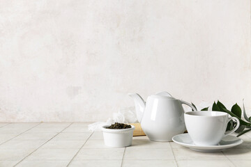 Teapot, cup of tea and bowl with dry leaves on white tile background