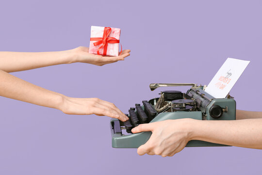 Female hands with gift box, retro typewriter and greeting card for International Women's Day on lilac background, closeup