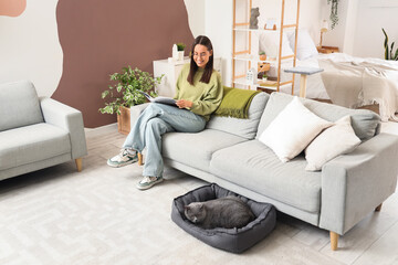 Obraz premium Young woman reading magazine and cute British Shorthair cat in pet bed at home