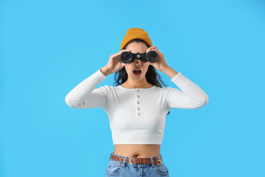 Shocked young African-American woman looking through binoculars on blue background