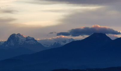 Fototapeta na wymiar Le cime delle montagne innevate al tramonto nel cielo plumbeo invernale e nuvole bordate di rosso dalla luce del sole