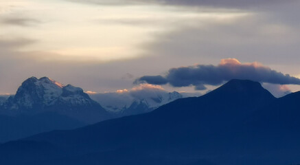 Le cime delle montagne innevate al tramonto nel cielo plumbeo invernale e nuvole bordate di rosso dalla luce del sole