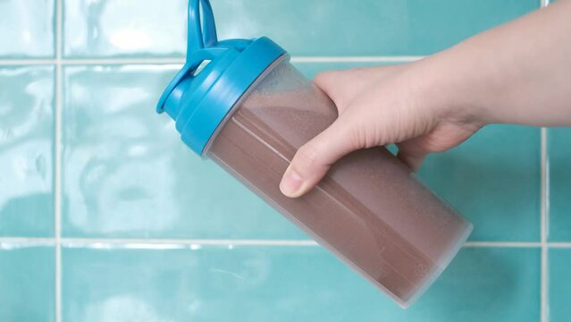 Female hand holds shaker with protein shake against background of mint tiles.