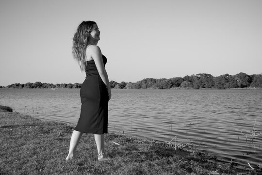 black dress, Latina with blue sky, and posing next to the lake, full body, face, posing at the dock, smiling in black and white
