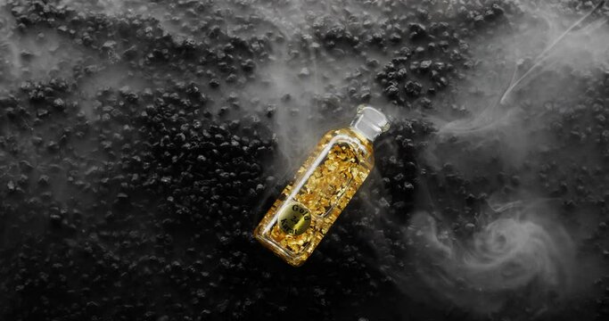 Bottle of 24k Gold falls into sea of smoke on a bed of black rocks