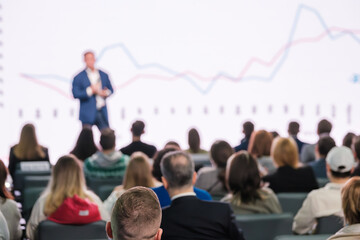 Blurry audience watching a presenter at a business conference with a financial growth chart in...