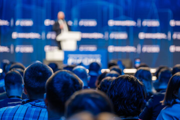 Blurred speaker presenting to an attentive audience at a professional corporate conference event.