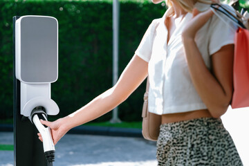 Young woman travel with EV electric car charging in green sustainable city outdoor garden in...