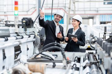 Asian female engineer and European male operator of an industrial plant discuss work and point fingers at steel and plastic production machinery, have laptops and walkie-talkies Wear a suit and helmet
