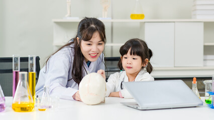 Asian woman doctor physiotherapist explains human fake skeleton skull model on a table to little cute children girl at laboratory study room. Education anatomical human concept learning for kids.