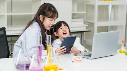 Asian woman scientist and little children girl reading laptop and tablet computer for data learning at chemical laboratory study room. Education research and development concept learning for kids.