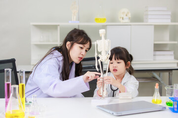 Asian woman doctor physiotherapist explains human fake skeleton hand model on a table to little cute children girl at laboratory study room. Education anatomical human concept learning for kids.
