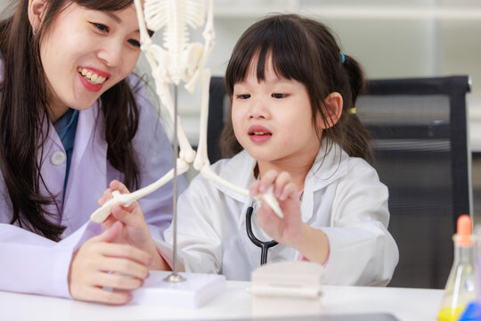 Asian woman doctor physiotherapist explains human fake skeleton hand model on a table to little cute children girl at laboratory study room. Education anatomical human concept learning for kids.