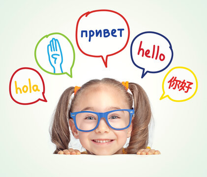 Little girl with eyeglasses leaning on table and looking at hello word in Spanish, English, Russian, Chinese and American sign language in speech balloons.