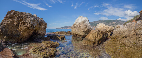Panoramic view of the rocky coast of the Mediterranean Sea with crystal clear water and between the stones you can see the silhouette in the distance of a sailing ship with lowered sails in Alanya.