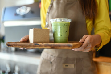 Young woman waiter in apron holding tray with Iced matcha latte to serve customer