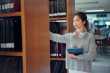 Young asian female college student taking book from shelf in library.