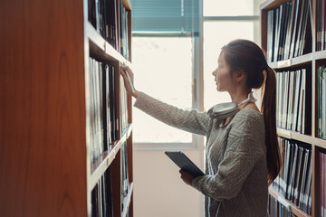 Young asian female college student taking book from shelf in library. - obrazy, fototapety, plakaty