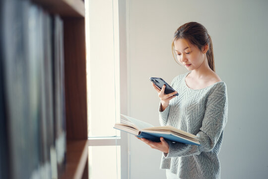 Young asian female college student using a smartphone taking a photo of a book page in the school library.