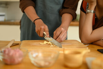 Close up of man in apron chopping garlic on wooden board while preparing meal with his wife in the kitchen