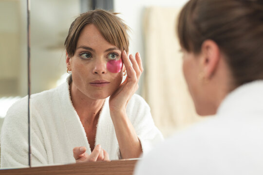 Caucasian woman examines her face in the mirror, applying a pink eye mask