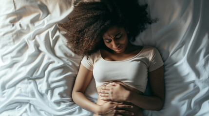 Overhead view of a black woman lying in bed, hands on stomach, subtle expression of pain, focus on personal healthcare