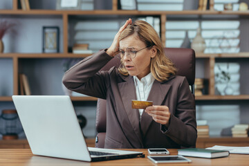 Worried mature businesswoman working at her office desk, dealing with payment problems using her credit card.
