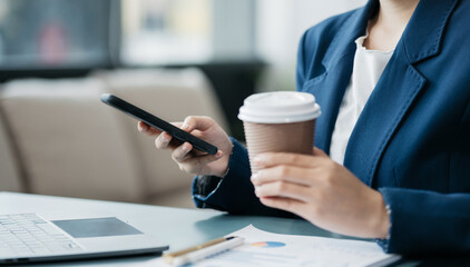 Asian businesswoman sits in a office working on laptop and enjoys a coffee.