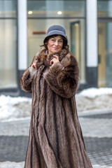 A stylish middle-aged woman in a fur coat and hat on a city street during the Christmas holidays