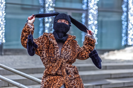 A stylish young woman in a leopard print fur coat and a balaclava with rabbit ears on a city street during the Christmas holidays