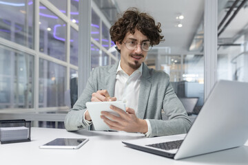 Fototapeta premium Professional young male with curly hair working at laptop in a corporate office setting, showing focus and expertise.