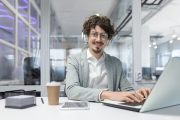 Curly-haired young mature businessman in a smart casual attire working diligently at his office desk with laptop and headset.