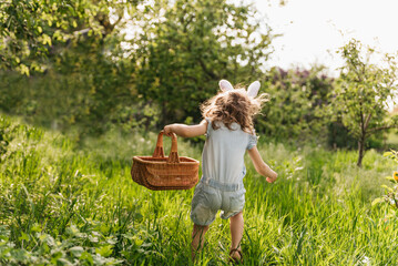 Easter egg hunt. Girl child Wearing Bunny Ears Running To Pick Up Egg In Garden. Easter tradition. Baby with basket full of colorful eggs. - 737858269