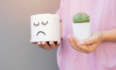 Sad face tissue roll and cactus holding by the hand of a young woman over light gray background.