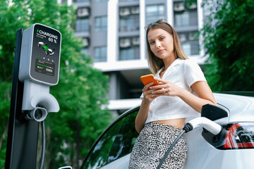 Young woman travel with EV electric car charging in green sustainable city outdoor garden in...