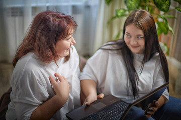 Fat funny funny adult mother and daughter with laptop indoors. A teenage girl teaches middle-aged woman modern technology. Internet, chatting